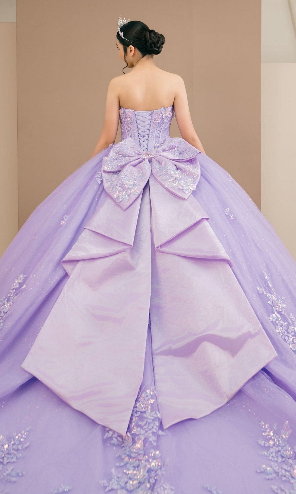 Wearing the Quinceanera Dress 8585JQ by Cinderella Couture—a strapless lavender ball gown adorned with floral details and a large bow at the back—a person stands with their back to the camera.